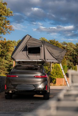Sahil yolunda park edilmiş gri lüks bir SUV, sert kabuklu bir çatı üstü çadırı açık ve merdivenle aşağı iniyor. Sıcak ışık ağaçlara vurur, bulutlar görünür, kamp havası ve hareket kabiliyeti.