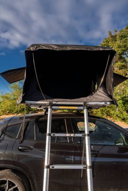Çatısı çadır ve merdiveni olan siyah lüks bir SUV park halinde. Parlak gün ışığı ve mavi gökyüzü kıyı parkı manzarası olduğunu gösteriyor. Geniş çadır flapları hızlı kurulumu gösterir.