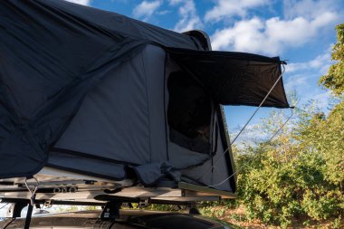 Dark gray rooftop tent on a luxury SUV, side window open with awning and guy lines, coastal shrubs nearby, late afternoon light, close up angled view, premium finish