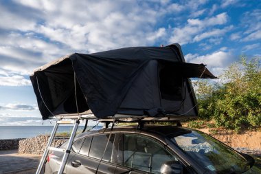 Çatısı çadır ve merdiveni olan koyu renkli bir SUV alçak taş duvarlar ve deniz kenarında kumlu bir çalılık, geniş açı, öğleden sonra ışığı, rüzgarlı bulutlar, seyahat havası.