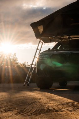 Çatıda çadırı olan lüks bir SUV kıyı şeridine yakın park edilmiş. Merdiven ön tekerleğin yanında duruyor, uzun gölgeler kum tonunda asfalt üzerinde uzanıyor, ve sıcak ışık alçaktan parlıyor..