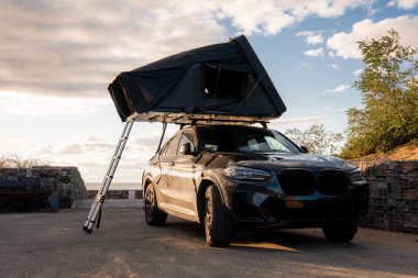 Kıyı gözcüsünün yanındaki karanlık lüks SUV parkları, alüminyum merdiven, alçak taş duvarlar ve ufukta ağaçlar, su ve bulutlar var.