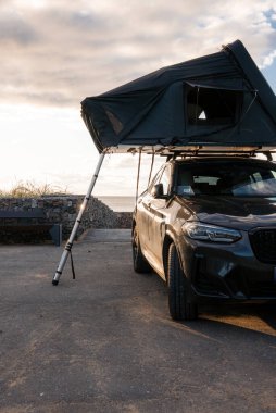 Gün batımında kayalık bir sahil gözcüsünün yanına park edilmiş koyu renkli lüks bir SUV merdiven bacaklarının üzerine kurulmuş bir çatı üstü çadırıyla. Ilık düşük ışık parlak panelleri ve çadır açılarını yansıtır.