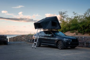 Siyah çatısı, çerçeve çadırı ve merdiveni olan koyu renkli lüks bir SUV, sahil kenarına park eder. Taş duvarlar, kumlu bitkiler ve okyanus ufku yumuşak altın saat ışığı altında belirir..
