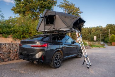 Koyu gri renkli lüks bir SUV, çatısı ve merdiveni olan Gabion taşından bir duvarın yanına park ediyor. Kıyıdaki bir katılımın arkasında çam ağaçları, kıyı şeridi yolu ve okyanus gece yarısı aydınlığında.