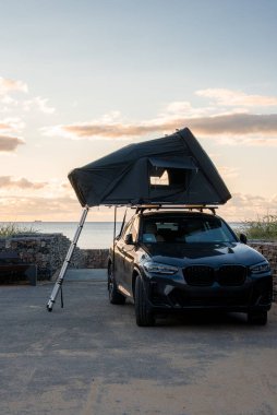 Çatı ve merdiveni olan karanlık lüks bir SUV gün batımında deniz kenarında, sakin bir okyanus ufkuna, dikey bir çerçeveye, yumuşak altın rengi parlak bir finişe bakıyor..