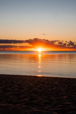 The sun touches the horizon and casts a golden vertical reflection on calm water, with a dark sandy beach in front and low clouds lit orange and amber in a vertical frame.