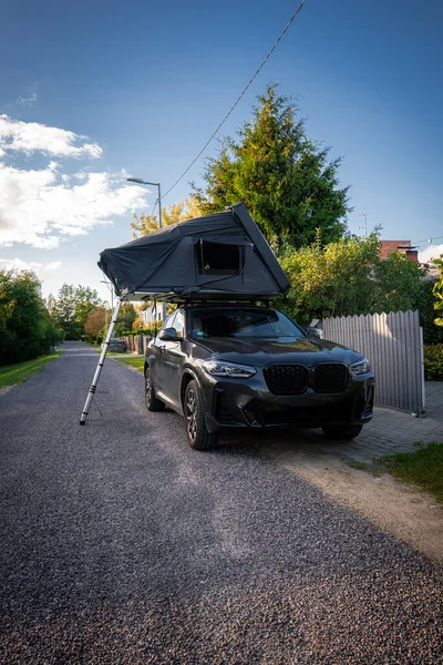 İkili böbrek ızgarası olan siyah lüks bir SUV, öğleden sonra aydınlığında merdiven, çit ve beyaz çitlerle döşenmiş bir çatı katı yolunda park halinde..