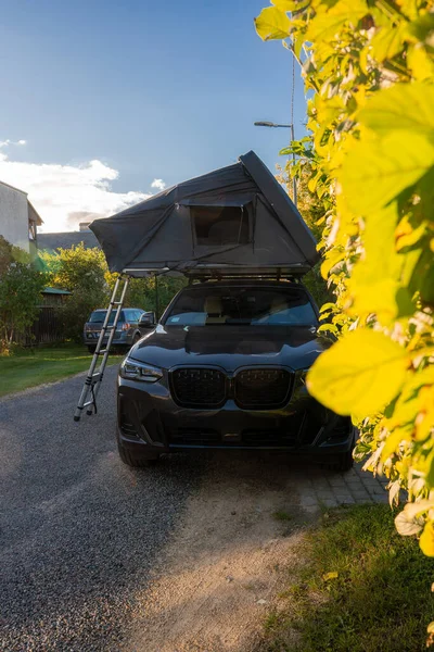 Güneş batarken banliyödeki evlerin yakınındaki sarı çitin kenarındaki dar çakıllı yolda çatısı ve merdiveni olan siyah lüks bir SUV, sıcak ışık ve sığ bir tarla derinliği