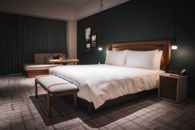 Japonya 'da modern bir otel odası beyaz çarşaflı bir yatak, bronzlaşmış bir yatak başlığı, kolye ışıkları ve kanepe ve masalı bir dinlenme odası gösteriyor. Yeşil aksanlı duvar ve sıcak ışıklandırma sakin bir ton oluşturdu.