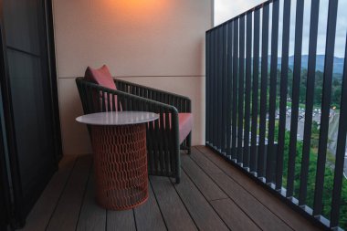 A high floor hotel balcony in Japan shows a cushioned lounge chair and lattice base side table, vertical slat railing, distant hills, and soft dusk lighting in a minimalist style.