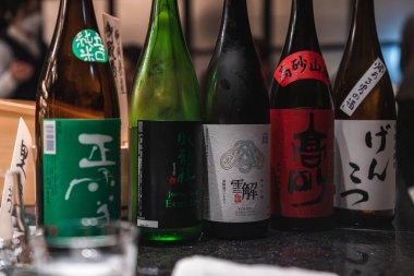 Kalın kanji etiketli beş sake şişesi Japonya 'da parlak bir barda duruyor. Yumuşak ortam ışığı ve bulanık patronlar yakın mesafeden küratörlü bir tatma sahnesi oluşturdular..