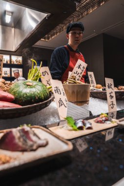 Japon bir aşçı, sezonluk malzemeler, saşimi tabakları ve açık mutfak teknesi ve tazeliği ön plana çıkaran sıcak ızgara ile parlayan parlak bir tezgahta durur..