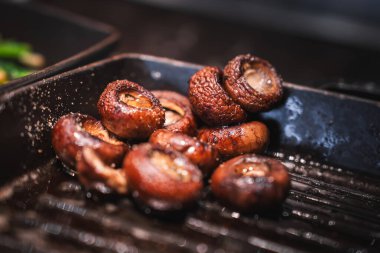 Small brown mushrooms sizzle on a ridged grill pan, caps seared with oil and coarse salt. Warm directional light catches steam and char in a tight shallow focus view.