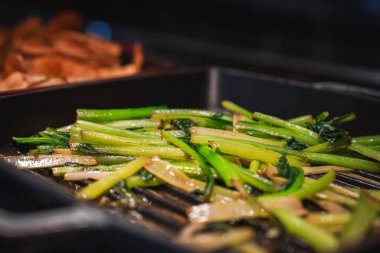 Close up of glossy green stems sizzling on a ridged grill pan in Japan. Light catches char and steam, with browned root slices softly blurred in the background.
