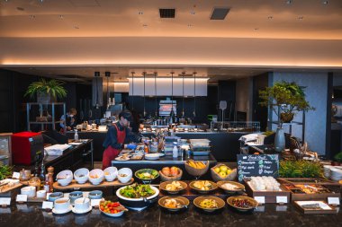 Japonya 'da bir otel büfesi siyah tezgahta salata, çorba, ekmek ve sıcak yemek sunar. Kırmızı önlüklü bir şef açık mutfakta kolye ışıklarıyla çalışır..