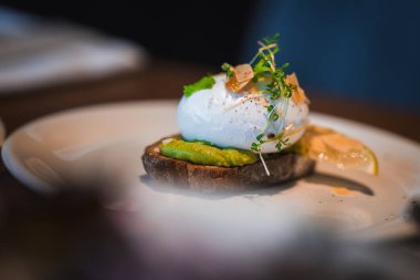 Yakından bakınca avokado tostu, haşlanmış yumurta, mikro yeşillikler, tıraş edilmiş peynir ve rafine edilmiş bir Japon restoranında sıcak ışık ve sığ odaklı bir limon dilimi görülüyor..