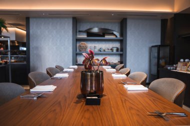 Uzun tahta bir masa beyaz peçeteler, yemek çubukları, pembe bromeliad süslemeleri ve sıcak, modern bir iç mekanda tabak ve yemek takımlarıyla dolu raflarla kurulur..