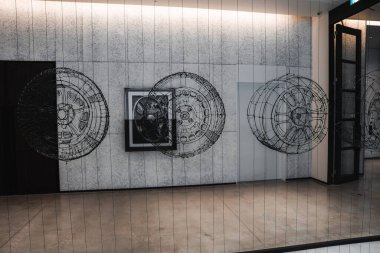 Minimalist bir koridorda, merkezde çerçevelenmiş gri bir duvarda otomotiv tekerleği çizgisi sanatı sergileniyor. Parlak taş zemin, cam kapı sağa, tenha ışıklar daha da parlıyor..