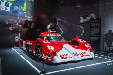 Kırmızı ve beyaz Toyota GT 1 TS020, Le Mans tabelası ve galeri ışıklandırması ile birlikte kokpit ve bileşenleri göstermek için kaldırılan bir müzenin halatlı platformunda duruyor..