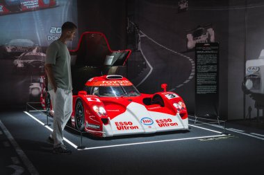 3 numaralı kırmızı ve beyaz bir Toyota TS020 GT 1 ışıklı bir platformda durur, Esso Ultron ve Toyota logoları görünür, ziyaretçi gözlemleme, pankartlar ve yarış fotoğrafları duvarları.