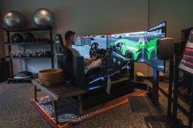 Bir adam kova koltuklu bir yarış simülatörü kullanıyor, güç geribildirim tekerleği, ve yeşil bir GT otomobili gösteren üç kavisli monitörler, bir otel fitness odasında, raflarda teçhizatla.