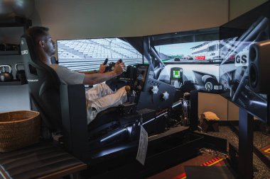 Yarış simülatöründe oturan bir adam kova koltuğu, tekerlek, pedallar ve önünde tribünleri ve arabaları olan yarış pisti manzarasını gösteren üç kıvrımlı monitörlerle.