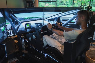 Bir adam, modern bir Japon otel salonunda üç ekran, tekerlek, pedal ve vitesli yarış simülatörü kullanıyor. Devrede kokpit manzaralı, yumuşak doğal ışık ve teknolojik odaklı..