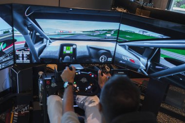 Bir kişi yarış simülatörü teçhizatı kullanır, üç ekran üzerindeki bir GT kokpiti gibi sürükleyici tekerlek ve pedallar sağ eğriye tribünlerle yaklaşır, HUD vites, hız ve turları gösterir..