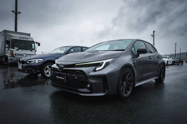Matte gri Toyota Corolla GR Sport ıslak bir çayırda KRM kamyonu ve koyu renkli sedan ile oturuyor, nemli asfalt bir Japon yarış tesisindeki bulutlu ışık altında yansıyor..