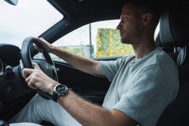 Bir adam modern bir Lexus 'ta oturuyor, elleri 9' da ve 3 'te, yan cama yağmur yağıyor. Bulanık yeşil ve sarı bariyerler Japonya 'da bir yarış pisti ya da test pisti olduğunu gösteriyor..