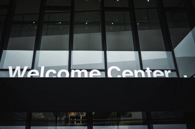 Low angle view of a glass facade with Welcome Center lettering under an overhang, reflective panels and dark metal frame suggest a public venue with soft interior lighting.