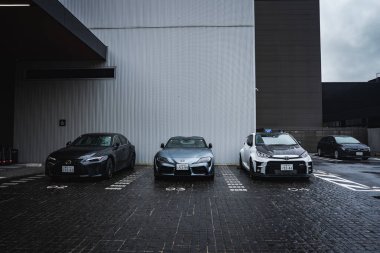 Modern bir otelin dışında üç Toyota aracı Japonya 'da yan yana park ediyor. Islak kaldırım taşları gri bir GR Supra, koyu renkli bir coupe ve mavi aksanlı beyaz bir minivan yansıtıyor..