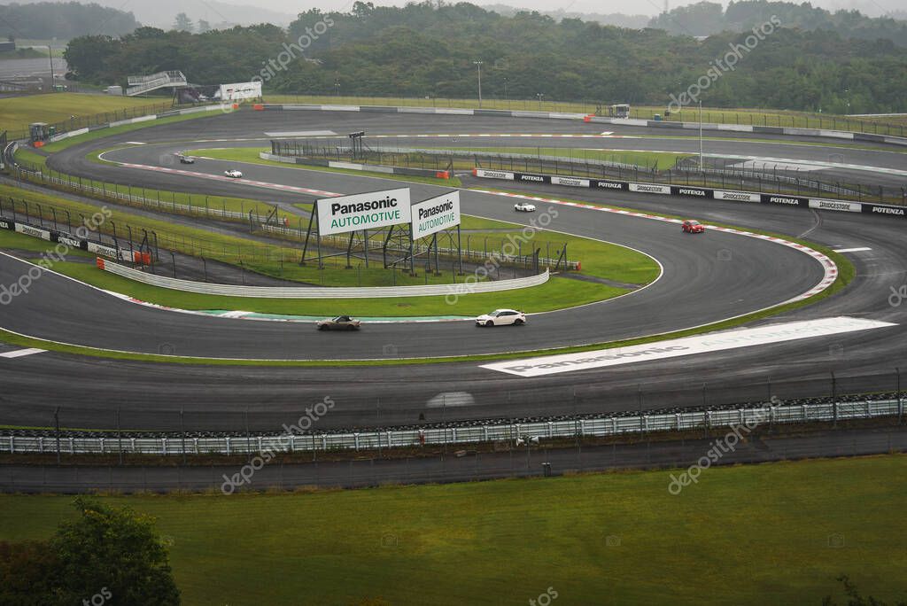 Sports cars navigate wet bends and chicanes at Fuji Speedway, Japan. Rain slick asphalt, green runoff, and Panasonic Automotive signs define a layered, wide view.