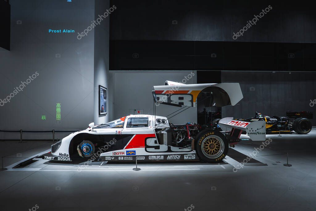 A vintage Group C style race car sits in a dim museum gallery in Japan, rear bodywork open to show engine, suspension, and gold wheel, number 9 and rear wing visible.