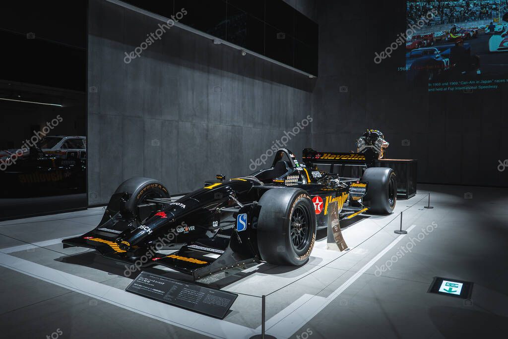 A black open wheel race car with yellow accents sits on a raised platform in a Japanese automobile museum, with velvet stanchions, placards, and a pit crew mannequin.