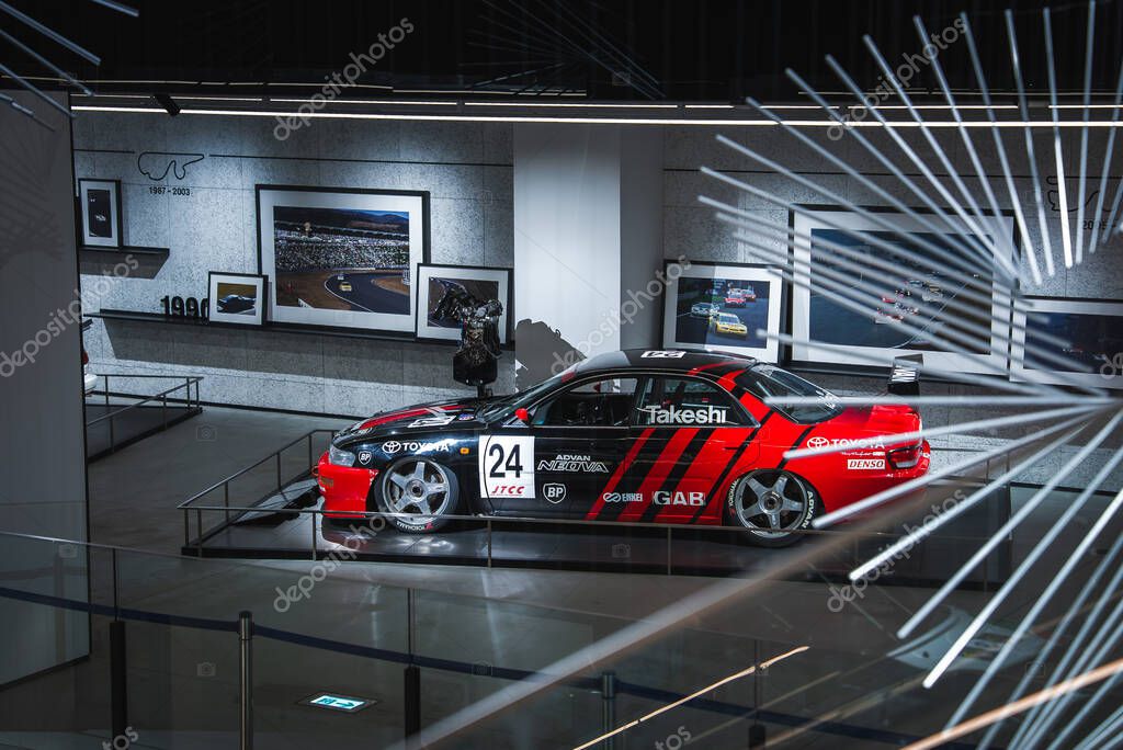 A red and black Toyota race car marked 24 and Takeshi sits on a raised platform with glass railings in a Japanese automotive museum under crisp gallery lighting.