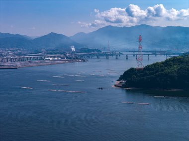Hiroşima Körfezi 'nin havadan görünüşü, önplanda Miyajima, mavi sulardaki istiridye çiftlikleri, uzun köprüler, kırmızı şanzıman kulesi ve yumuşak öğleden sonra ışığı altında sanayi alanları..
