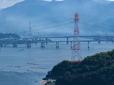 Japonya 'nın Hiroşima yakınlarındaki hava manzarası geniş bir kanal üzerinde köprüler, kırmızı ve gümüş şanzıman kulesi, yüksek gerilim hatları ve sisli tepeleri olan su ürünleri salları gösteriyor..