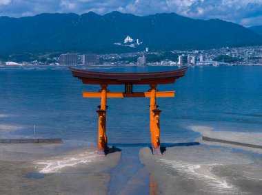 Turuncu torii, Japonya 'nın Hiroşima kentindeki mavi bir körfezin önünde gelgit düzlüklerinde duruyor. Merkezli kompozisyon, açık gün ışığı, temel görünür yansıma dalgalanmaları, şehir ve dağlar mevcut..