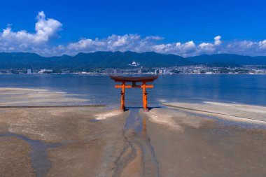 Hava görüntüsü, Japonya 'nın Miyajima şehrinde gelgit düzlüklerinde Büyük Torii' yi gösteriyor. Hiroşima Körfezi parıldıyor, şehir ve mavi dağlar gün ortasında ufku kaplıyor..