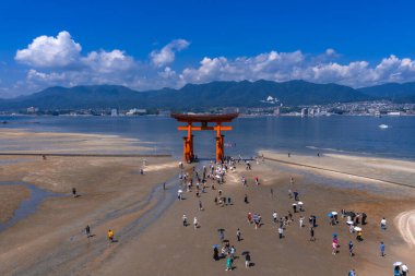 Hava görüntüsü, Japonya 'nın Hiroşima yakınlarındaki Miyajima' da, Büyük Torii 'ye yürüyen insanları gösteriyor. Gün ortası ışığı, geniş açı, mavi gökyüzü, deniz, dağlar ve şehir suyun ötesinde..