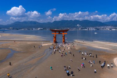 Japonya, Hiroşima yakınlarındaki Itsukushima Tapınağı 'ndaki Büyük Torii' deki gelgit düzlüklerinde yürüyen insanların hava görüntüsü. Dağları, şehri ve sakin Seto İç Denizi..