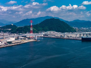 Japonya, Hiroşima 'nın hava görüntüsü, koyu mavi bir liman üzerinde beyaz bir kemer köprüsü, kırmızı bir şanzıman kulesi, rıhtım, şehir limanı, tepe ve öğlen vakti uzak dağları gösteriyor..