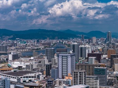 Hava görüntüsü, Hiroşima 'nın merkezini gün ışığında, birden fazla köprüsü olan geniş bir nehir ve kümülüs bulutlarının altında mavi yeşil dağlar gösteriyor..