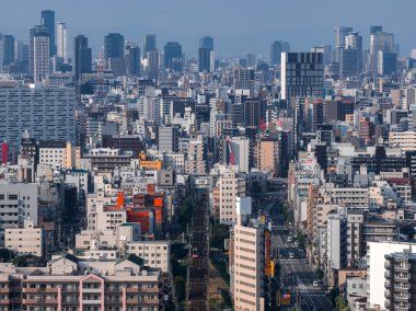 Osaka, Japonya şehir manzarası orta bloklar, demiryolu hatları ve geniş bir cadde gösteriyor. Turuncu aksanlar gri ve beyaz cepheleri gösteriyor. Bulanık gün ışığı katmanlı derinliği ve ölçeği ortaya çıkarır..