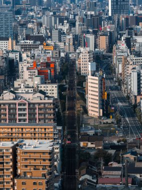 Japonya, Osaka 'nın hava manzarası, yoğun bloklar, ofis kuleleri ve merkezi bir demiryolu koridorunu gösteriyor. Serin gün ışığı pastel cephelerde ve çok şeritli yollarda uzun gölgeler bırakır..