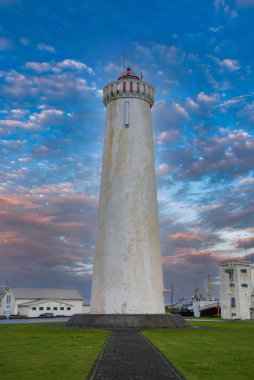 Gün batımında İzlanda 'da çimenli bir güvertede kırmızı fenerli beyaz bir fener duruyor. Düz bir patika, liman direkleri ve sanayisi ile üsse gidiyor..