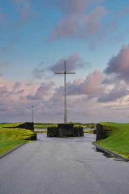 Alacakaranlıkta İzlanda kıyı mezarlığının yanındaki taş platformda uzun metal bir haç duruyor, ıslak kaldırımlar ve su birikintileri pastel bulutları yansıtıyor, lav kaya duvarları manzarayı sınırlıyor..