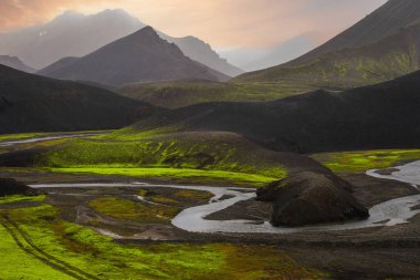 Sarmal bir nehir, Landmannalaugar, İzlanda yakınlarındaki siyah volkanik tepeleri ve yosun ovalarını şafak veya günbatımında, ılık ışık ve sisli katmanlı dağları keser..